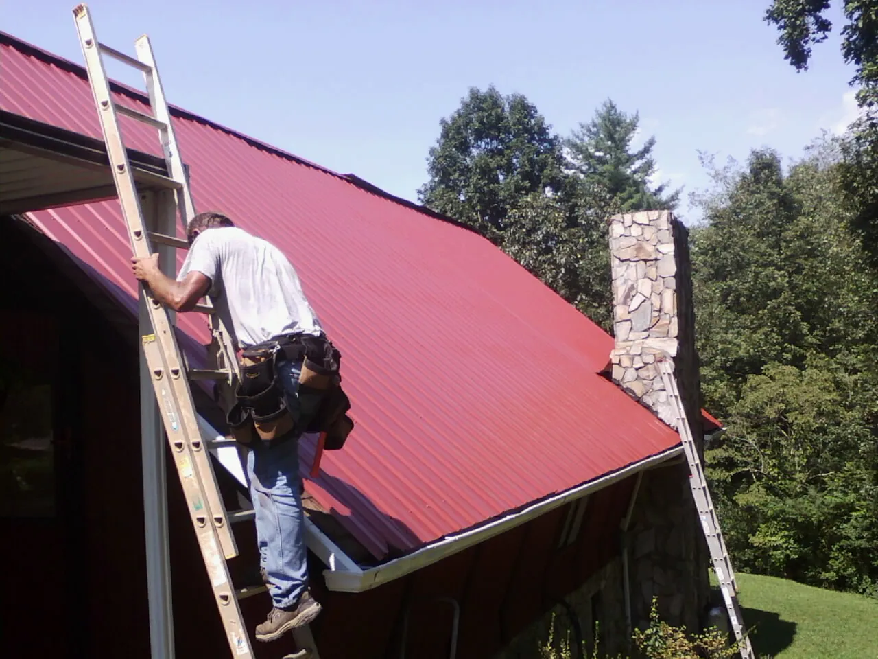Roofer on a ladder beside a red metal roof and stone chimney — Soffit & Fascia Repair by Roofing Brothers in Tompkinsville